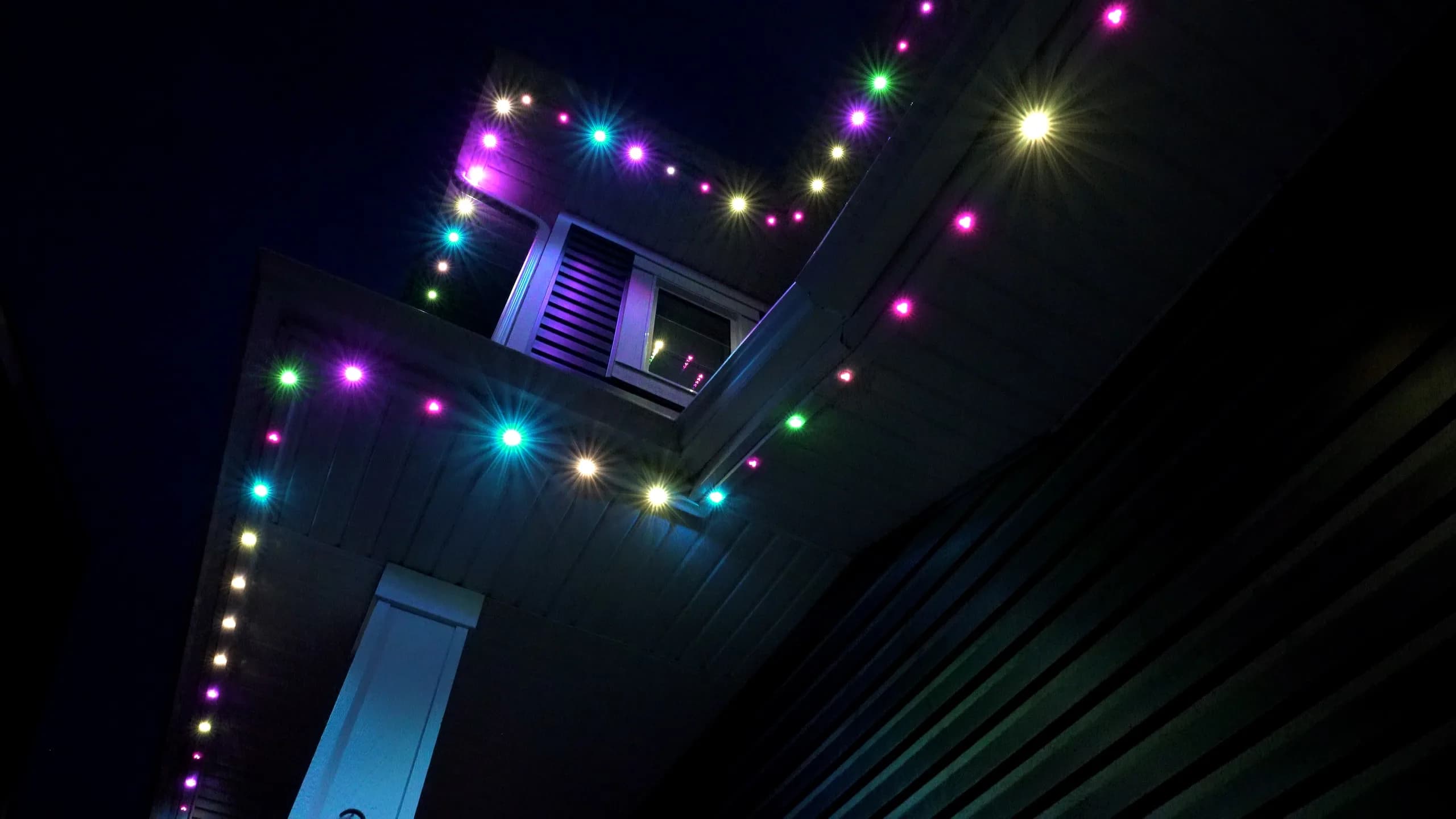 Close view of a home's soffit and eaves at night, individual Lumenary Pro emitters picked out in pink, green, cyan and warm white against a black sky.